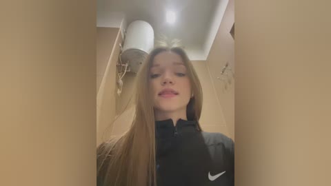 A video of a young, light-skinned girl with long, straight blonde hair, wearing a black Nike jacket, standing in a beige-walled hallway with a white cylindrical light fixture on the ceiling.