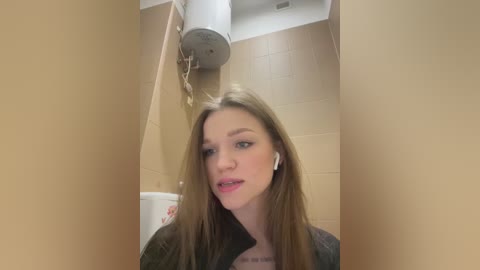 Media: A video of a young woman with straight, long brown hair, light skin, and blue eyes, wearing a black jacket and white earphones, standing in a beige-tiled bathroom with a white toilet and a metal heater.