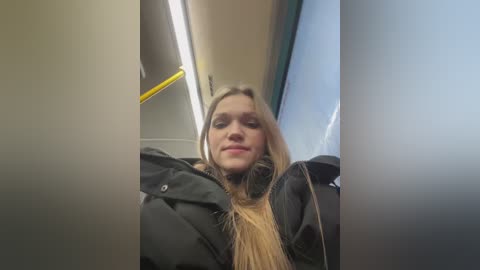 Media: Video of a young Caucasian woman with long blonde hair, wearing a black puffer jacket, smiling slightly, taken inside a train. The background shows the train interior with yellow handles and fluorescent lights.