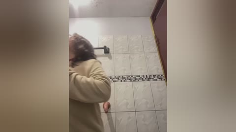 Media: Video of a person with shoulder-length brown hair, wearing a beige sweater, standing in a bathroom with white and black tiled walls and a black-and-white mosaic pattern. The image is taken from a slightly tilted angle, showing the person's back and upper body.