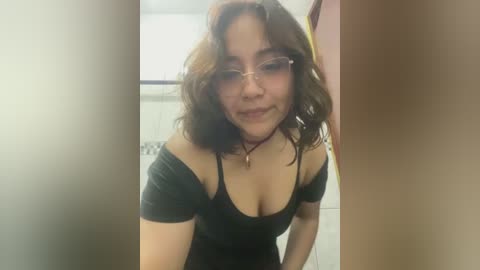 Media: Video of a smiling young woman with medium brown skin and curly hair, wearing a black off-shoulder top, standing in a bathroom with white tiles.