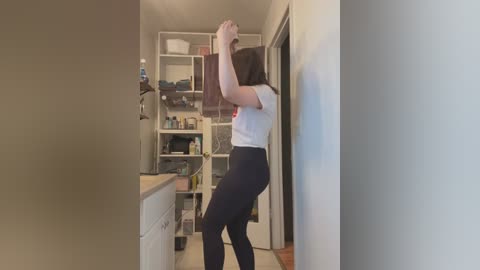 Media: Video of a woman in a white t-shirt and black leggings stretching against a white wall in a narrow, cluttered kitchen.