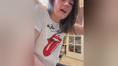 A video of a woman with shoulder-length brown hair, wearing a white t-shirt with a red tongue graphic and black text. She has a slightly open mouth, appearing surprised. Background includes a white cupboard and beige tiled floor.