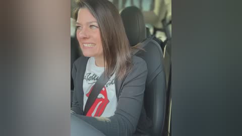 Video of a smiling, light-skinned woman with shoulder-length brown hair, wearing a grey cardigan over a white graphic tee, seated in a car with a seatbelt on, viewed through a window.