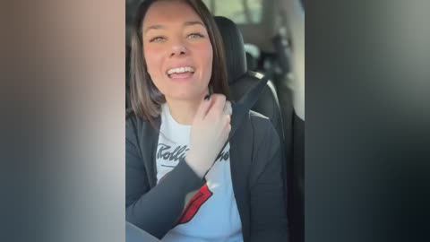 Media: A video of a smiling woman with shoulder-length brown hair, wearing a white T-shirt and black jacket, buckled in a car seat.