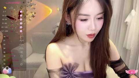 Media: Video of an East Asian woman with long brown hair and fair skin, wearing a strapless purple dress with a bow, indoors, looking down. Background features a white chair and white curtains.