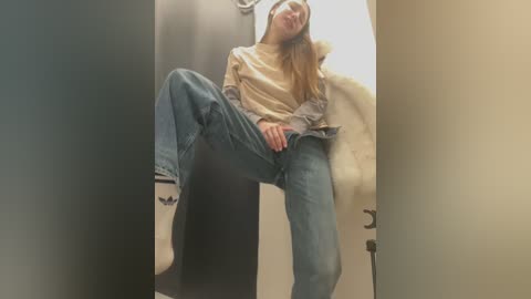 Video of a young, slender woman with long brown hair, wearing a beige shirt, gray jacket, blue jeans, and Adidas sneakers, sitting in a public restroom with legs spread, her face slightly blurred.