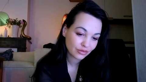 Media: Video of a fair-skinned woman with long black hair, wearing a black top, seated in a dimly lit living room with beige furniture, potted plants, and a warm ambiance.