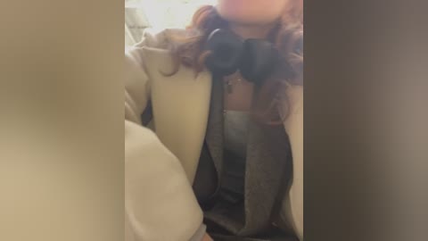 Media: Video of a woman with curly brown hair wearing a beige coat, grey sweater, and black headphones, leaning against a beige wall, blurred background.