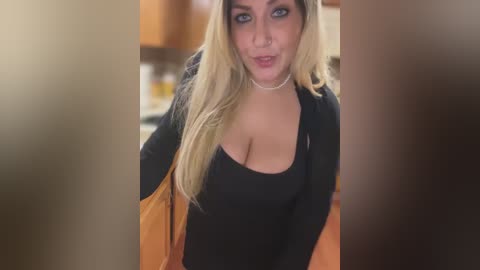 Media: A video of a young, fair-skinned woman with long, blonde hair, wearing a black top and a beige jacket, standing in a blurry kitchen.