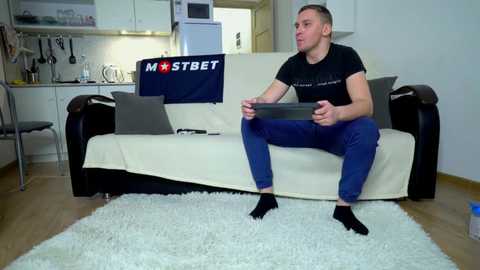 Media: Video of a young man with short hair, wearing a black t-shirt, blue jeans, and black socks, playing a video game on a couch in a modern, minimalistic living room.