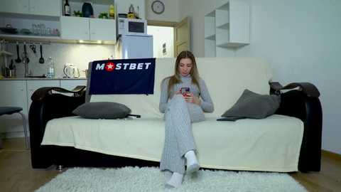 Media: Video of a young woman in gray pajamas, sitting on a white sofa with gray pillows, in a modern, minimalist living room with a kitchen in the background.