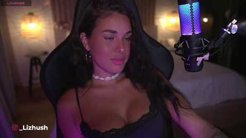 Video of a young woman with long dark hair, wearing a black lace bra and a white choker, sitting in a gaming chair under purple and blue studio lights.