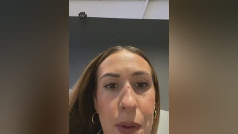 Media: A close-up video of a Caucasian woman with fair skin, long brown hair, and light makeup. She wears hoop earrings. The background shows a plain, gray wall with a ceiling light fixture.