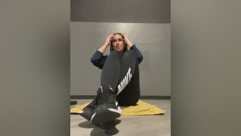 Media: Video of a woman sitting on a yellow mat in a dimly lit room, wearing black Nike sneakers and leggings, with hands on head, appearing distressed.