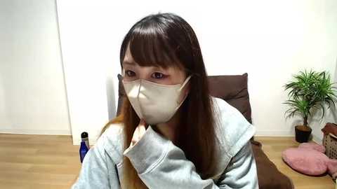 Media: Video of a young Asian woman with long brown hair, wearing a gray hoodie and white mask, sitting in a brown chair, in a minimalistic room with wooden floor, potted plants, and blue bottle.