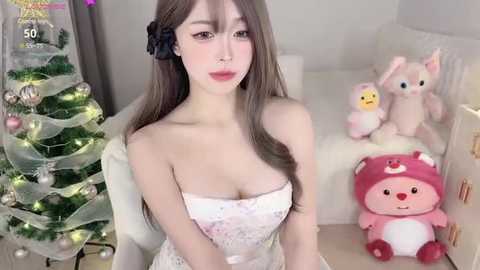 Media: Video of a fair-skinned, slender Asian woman with long brown hair, wearing a white strapless dress, standing next to a decorated Christmas tree and plush toys.