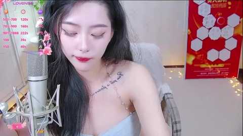 Media: Video of an Asian woman with long black hair, red lipstick, and a delicate tattoo on her shoulder, singing into a silver microphone in a cozy room with a red poster and colorful lights.