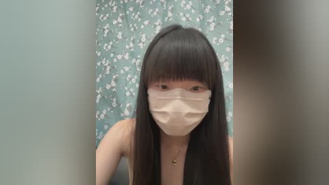 Media: Video of an Asian woman with long, straight black hair and bangs, wearing a beige surgical mask, seated indoors against a floral-patterned teal wall.