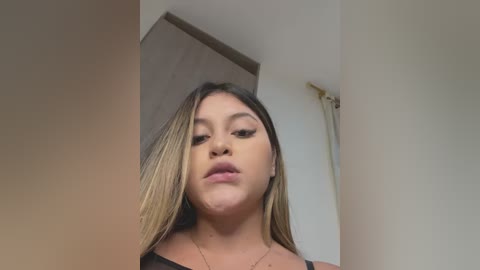 Media: Video of a Latina woman with long, straight, light brown hair, light skin, and full lips, wearing a black top, standing indoors against a beige wall.