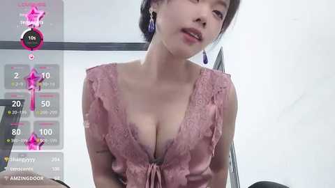 Media: Video of an Asian woman with fair skin and medium-length black hair in a pink lace top, leaning forward, in a bright room with a white wall.