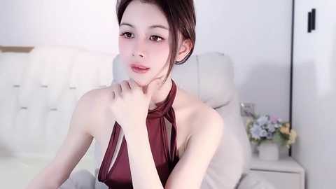 A video of an Asian woman with pale skin, dark hair, and light makeup, wearing a sleeveless, halter-neck maroon top, seated on a white couch in a minimalist, modern room.