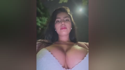 Media: Video of a young woman with long dark hair, wearing a white lace bra, capturing her ample cleavage. The background is dimly lit with blurred greenery.