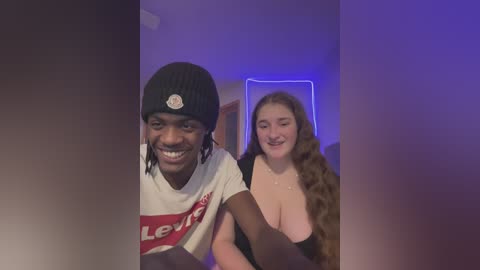 Video of a smiling, dark-skinned man with a black beanie and white t-shirt, and a smiling, light-skinned woman with long brown hair, wearing a low-cut black top, standing indoors under blue LED lights.