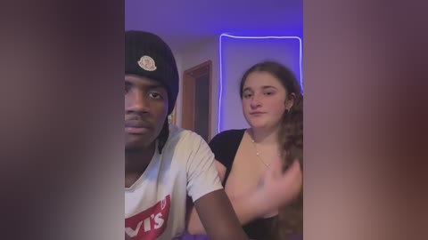 A video shows a young Black man in a white \"Vans\" t-shirt, black beanie, and a woman with long brown hair, both smiling, in a dimly-lit room with purple lights.