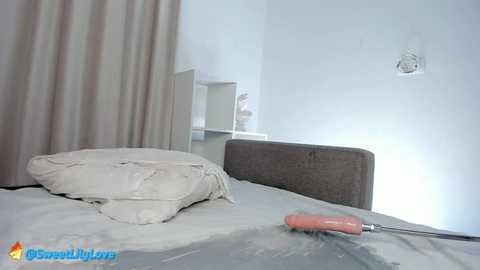 A minimalist, modern bedroom with a gray upholstered headboard, beige curtains, and a white wall-mounted shelf. A white pillow and a pink screwdriver lie on the bed.