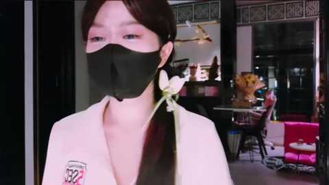 Media: A video of a woman in a black face mask, white shirt, and a white flower tucked behind her ear. The background shows an indoor space with dim lighting, a table, chairs, and decorative items.