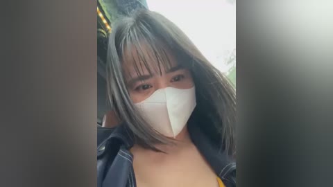 Media: Video of a young Asian woman with medium skin tone, straight black hair, and bangs, wearing a white surgical mask, blue denim jacket, and a yellow top. She appears to be in a dimly lit, possibly outdoor setting.