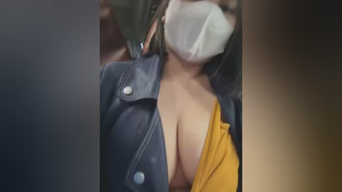 Media: Video of a woman with light brown skin, wearing a blue leather jacket, mustard-yellow shirt, and a white mask, partially revealing her ample cleavage. Background is blurred, indoor setting.