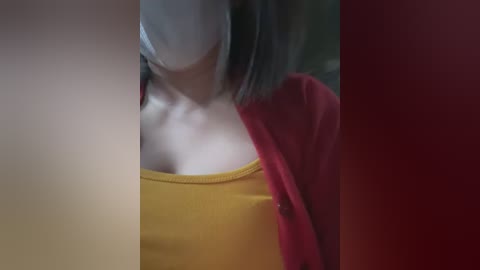 Media: Video of a woman with a pale complexion, wearing a yellow tank top, red cardigan, and a white mask, partially visible, blurred background.