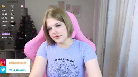 A video of a young, fair-skinned woman with straight, shoulder-length blonde hair, wearing a light blue t-shirt with \"MAKE A WISH\" text and a cartoon character. She sits in a pink gaming chair in a softly lit room with a Christmas tree in the background.