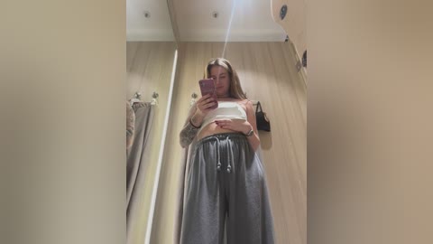 Media: Video of a tall, light-skinned woman with long brown hair, wearing a white crop top and loose gray sweatpants, taking a mirror selfie in a beige-walled dressing room.