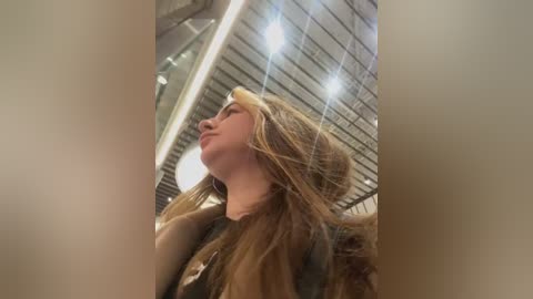 Media: A video of a young woman with long blonde hair, wearing a tan leather jacket, leaning against a glass wall under fluorescent lighting in a modern indoor setting.