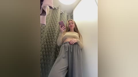 Media: Video of a young woman with long blonde hair, wearing a beige crop top and grey sweatpants, taking a selfie in a bathroom with a quilted green shower curtain.