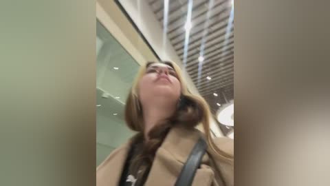 Media: A low-angle video of a woman with long blonde hair, wearing a beige coat, looking upwards. The background features a modern, indoor space with glass panels and a ceiling with horizontal slats.