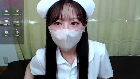 Media: Video of an Asian woman in a white nurse's uniform and cap, wearing a surgical mask, indoors with beige curtains in the background.