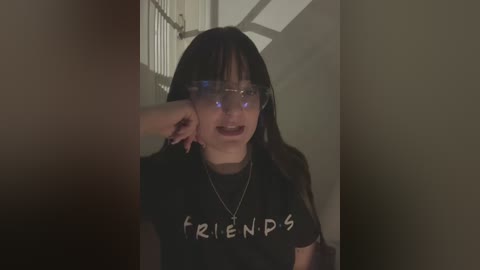 Media: Video of a young woman with long dark hair and pale skin, wearing black glasses and a \"Friends\" T-shirt, leaning against a dimly lit, narrow hallway with metal bars visible in the background.