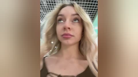 Media: Video of a fair-skinned woman with long blonde hair, wearing a black tank top, standing in a modern, tiled bathroom with a white grid ceiling.