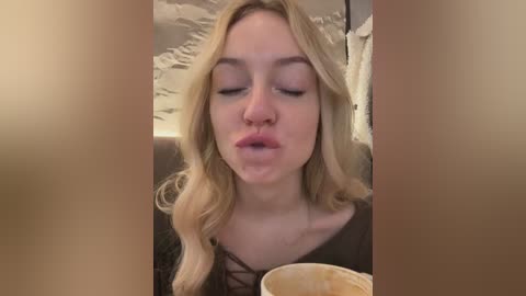 Media: A close-up video of a young Caucasian woman with long, wavy blonde hair, wearing a dark top, blowing a bubble with her lips. She sits indoors, with a beige wall and a white ceiling in the background.