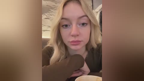 Media: Video of a young, fair-skinned, blonde woman with light makeup, wearing a dark top, seated at a table, holding a brown, cylindrical object, blurred background.