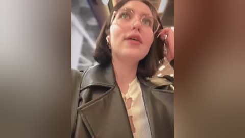 Media: Video of a young woman with shoulder-length brown hair, wearing glasses, a green leather jacket, and a white blouse, looking upward thoughtfully in a subway car.