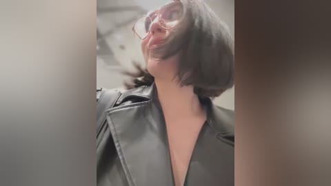 Media: A video of a woman with short black hair, wearing glasses, a black leather jacket, and a deep neckline, captured in an urban setting.