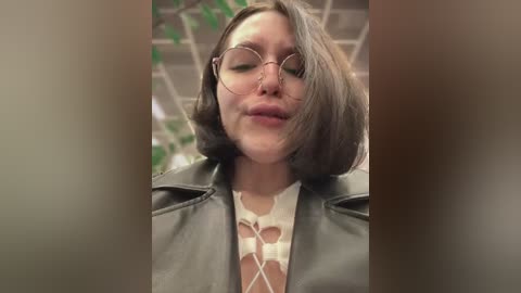 Media: A video of a young woman with short brown hair, wearing glasses, a black leather jacket, and a floral-patterned top, set against a blurred indoor background with green vines.