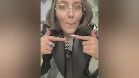 Media: Video of a young woman with short, wavy gray hair, wearing glasses, a black leather jacket, and a white shirt with a black skull pattern, pointing at the camera.