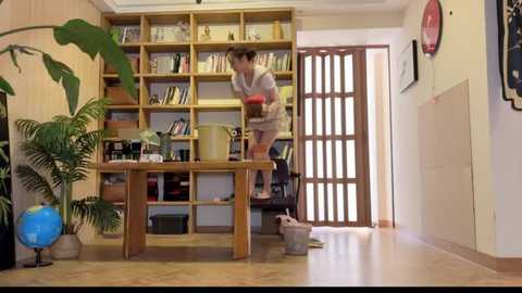 Media: Video of a woman with brown hair in a beige sweater and shorts, vacuuming a wooden floor, surrounded by bookshelves and a banana leaf plant.