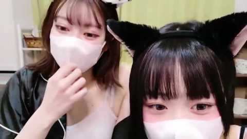 Media: Video of two young Asian women wearing white face masks and cat ears, with one holding a finger to her mouth, in a brightly lit room with green curtains.